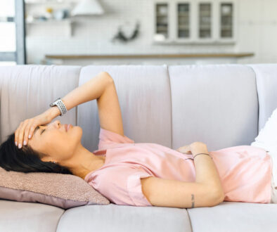 Young Asian Woman Laying On a Couch Holding Her Stomach and Head Experiencing Common Pregnancy Symptoms
